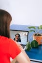 Woman in red shirt video chatting on laptop with plant nearby at home.